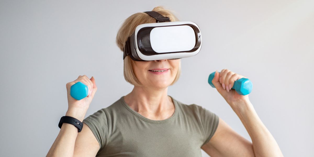 Mature woman training with dumbbells, wearing virtual reality headset, having domestic workout. Senior lady doing sports with VR application. Futuristic fitness and contemporary technologies
